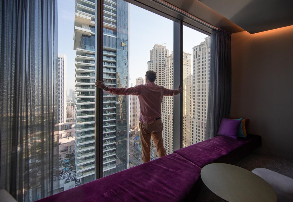 Person viewing Dubai skyline from luxury apartment window with purple bedding, showcasing Airbnb investment property potential